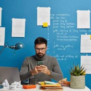 Busy bearded male programmer ponders on task, concentrated into smartphone, wears optical glasses, p